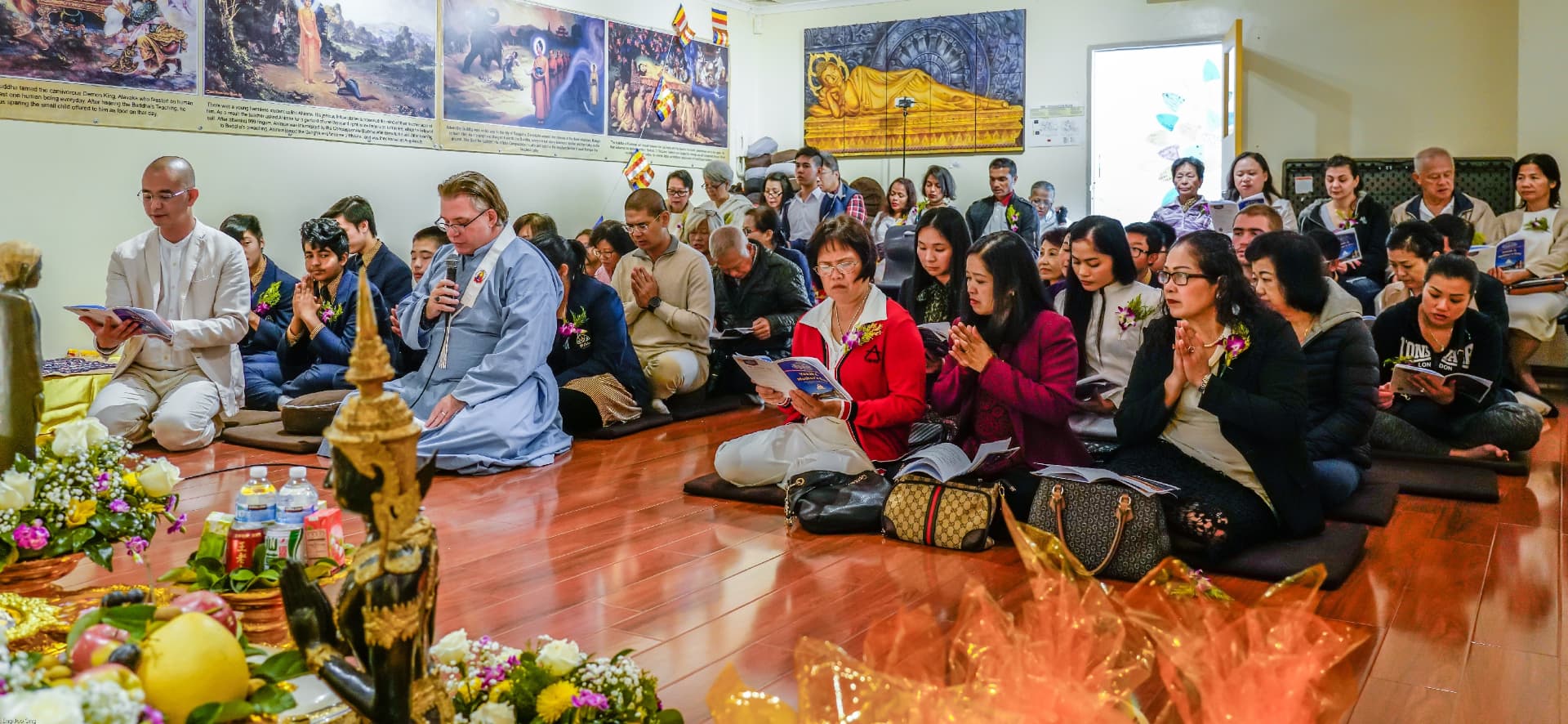 The community seated in the hall for Vesak, chanting books in hand, with offerings of flowers and fruit in the foreground